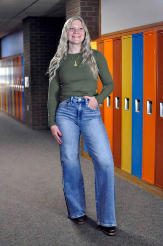 A woman stands in a school hallway wearing an olive green long-sleeve top and light-wash blue flare jeans, with colorful lockers in the background.