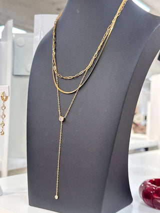 A gold layered necklace displayed on a black jewelry bust. The necklace features three distinct layers: a thick textured chain, a thin twisted chain, and a delicate chain with a small round clear stone pendant and a longer drop chain ending in a tiny bead.
