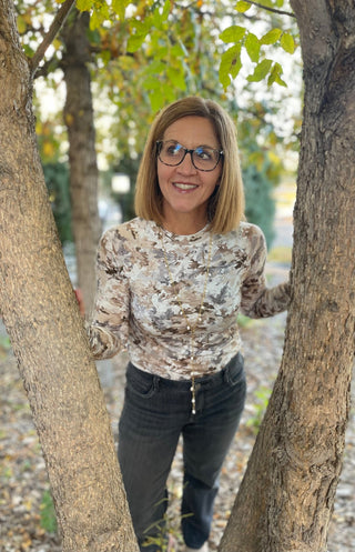 A woman with blonde hair and glasses stands outdoors between two tree trunks, wearing a beige and brown abstract patterned long-sleeve top and dark gray jeans.
