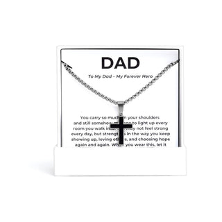 A silver cross pendant necklace displayed on a white gift card that reads 'DAD To My Dad - My Forever Hero' with a supportive message below, photographed on a clean white background.