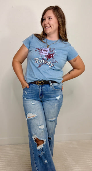 A woman modeling a light blue graphic t-shirt with 'Mama Rowdy' printed on the front, paired with distressed blue jeans, standing against a plain off-white wall.