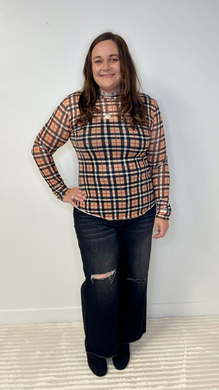 Person wearing a plaid shirt and jeans standing against a white wall.