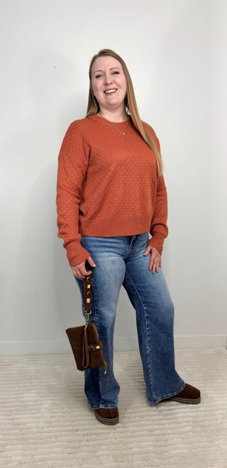 A woman modeling a textured rust-orange long-sleeve knit sweater, paired with blue flare jeans and brown shoes, holding a brown handbag against a plain white wall.