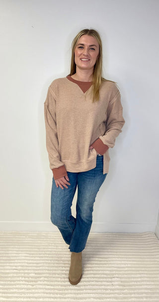 Woman wearing a beige sweater and blue jeans standing against a white wall.