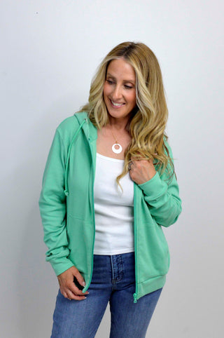 Woman wearing a green hoodie, white top, and blue jeans against a plain background