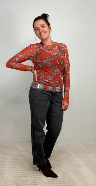 A woman modeling a rust-orange long-sleeve top with a blue and white paisley pattern, paired with dark gray jeans and dark brown boots, standing against a light gray wall.
