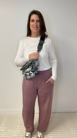 Woman wearing a white long-sleeve shirt and mauve pants, holding a camouflage-patterned bag against a white wall.