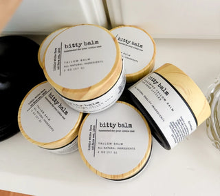 Five jars of bitty balm Tallow Balm arranged on a light surface. The jars have light wood-grain lids with white labels and black text, with one jar showing a pale yellow balm visible at the side.