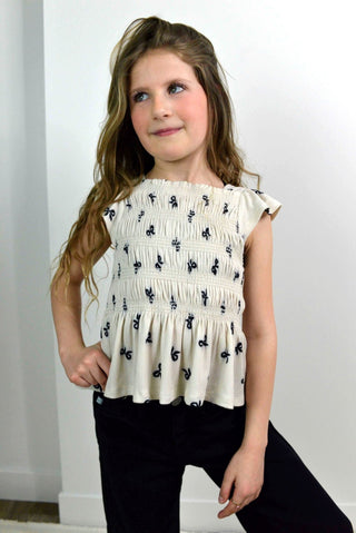Young girl wearing a white blouse with black patterns and black pants against a white background