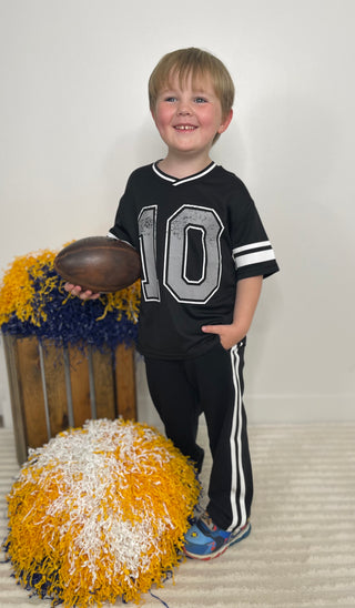 A young boy smiling and holding a brown football, wearing a black short-sleeve sports jersey with the number 10 on the front, black track pants with white side stripes, and blue sneakers. He is standing in a room with a white wall and beige carpet, next to yellow, blue, and white pom-poms.