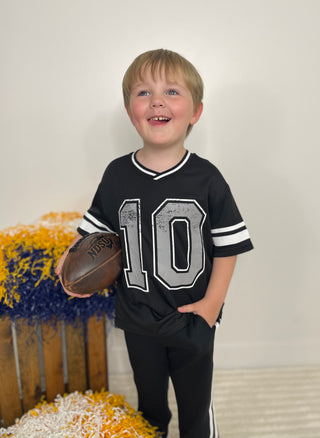 A young boy smiling and holding a football, wearing a black short-sleeve sports jersey with the number 10 in gray and white, paired with black pants. He is standing in front of a plain light wall with yellow and blue decorative pom-poms on the left.