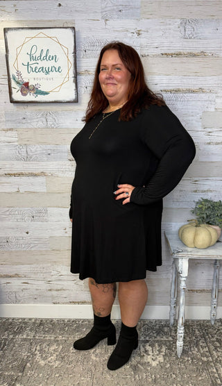 Woman wearing a black dress standing in front of a wooden wall with a sign that reads 'Hidden Treasure'.