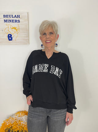 A woman with short gray hair and glasses stands wearing a black long-sleeve sweatshirt with 'GAME DAY' in white varsity-style lettering across the chest, paired with gray jeans. A wooden 'BEULAH MINERS' sign is visible on the wall behind her.