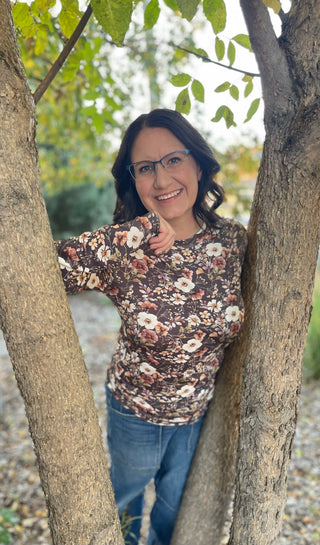 A woman with glasses and dark hair stands smiling between two tree trunks outdoors, wearing a brown floral long-sleeve top and blue jeans.