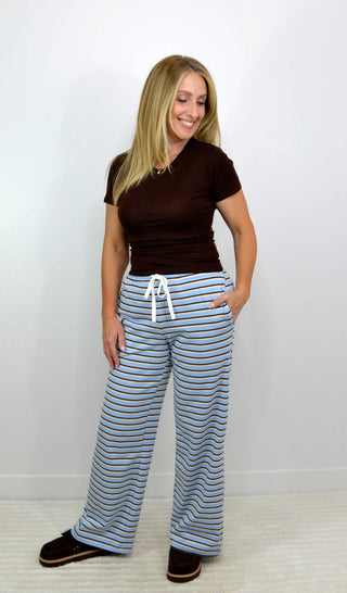 A woman modeling a pair of wide-leg lounge pants with blue and white horizontal stripes and a white drawstring, paired with a dark brown t-shirt and dark brown slippers, standing against a plain white wall.