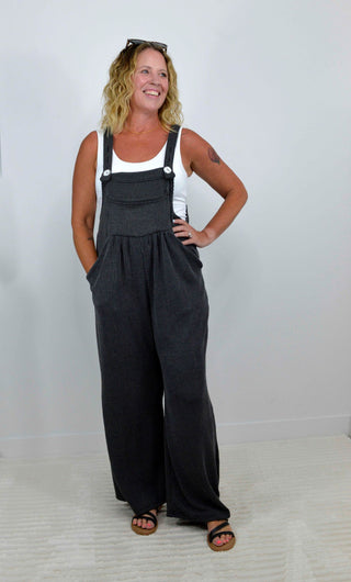 A woman modeling a pair of dark charcoal gray wide-leg overall jumpsuits over a white tank top, standing against a plain light gray wall.