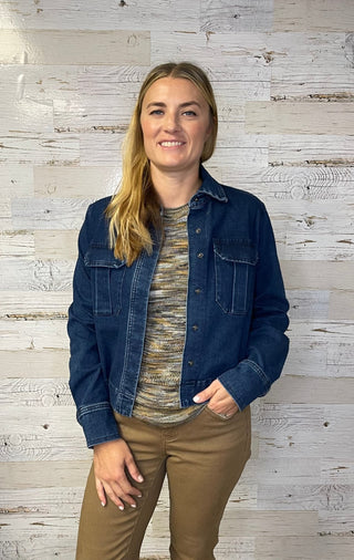 Woman wearing a denim jacket over a patterned shirt and brown pants against a textured wall.