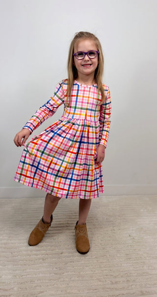 A young girl with glasses and brown boots stands in a room wearing a long-sleeve plaid dress with a multicolor pattern of pink, orange, yellow, green, and blue on a white base.