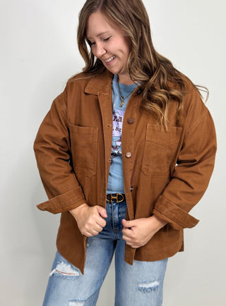 A woman wearing a brown button-up utility jacket over a blue graphic t-shirt and light-wash distressed jeans, standing against a plain light gray background.
