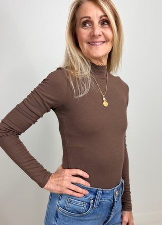 A smiling blonde woman wearing a fitted brown ribbed mock neck long-sleeve top and blue jeans, standing against a plain light gray wall.