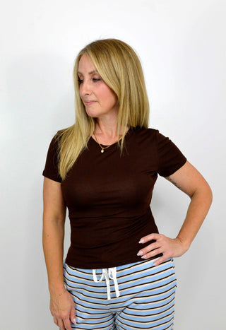 A woman modeling a fitted short-sleeve crewneck t-shirt in dark brown, paired with light blue and white striped pants, against a plain white background.