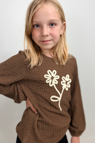 A young girl with blonde hair wearing a brown textured knit sweater with a cream-colored embroidered flower design on the front, photographed against a plain light gray background.