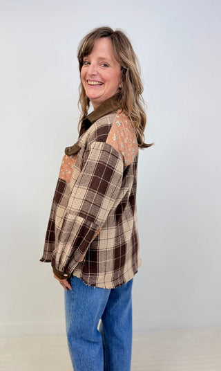 A woman modeling a brown and beige plaid patchwork flannel shirt with blue jeans, standing against a plain light gray studio background.