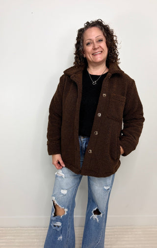 A woman with curly brown hair stands against a plain white wall wearing a dark brown sherpa fleece button-up jacket over a black top and distressed blue jeans.
