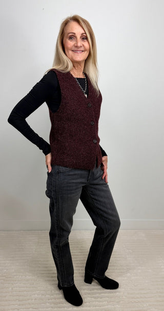 A woman modeling a dark burgundy knit button-up vest over a black long-sleeve shirt, paired with dark gray jeans and black shoes, standing against a plain light gray wall.