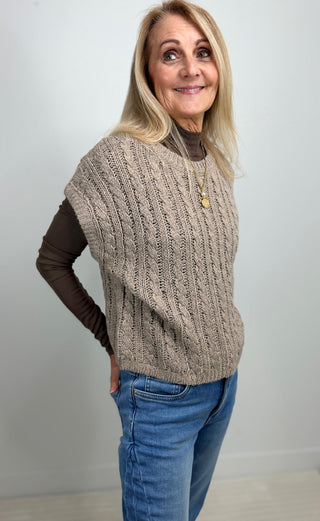 A woman modeling a beige cable-knit sleeveless sweater vest over a dark brown long-sleeve top and blue jeans, standing against a plain light gray wall.