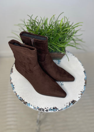 A pair of dark brown suede ankle boots with pointed toes, displayed on a white ceramic tray with a blue and black speckled rim, with a small green potted plant in the background.