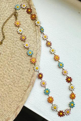 A colorful floral bead necklace with daisy-like flowers in white, yellow, orange, blue, and pink, draped over a straw hat on a white textured surface.