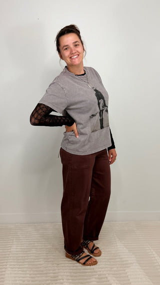 A woman modeling a gray graphic t-shirt with a black long-sleeve lace-up underarm layer, paired with dark brown pants and black sandals, standing against a light gray wall.