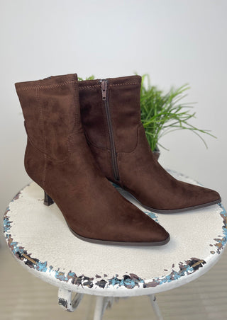 A pair of dark brown suede ankle boots with pointed toes and side zippers, displayed on a distressed white stool with a small green plant in the background.