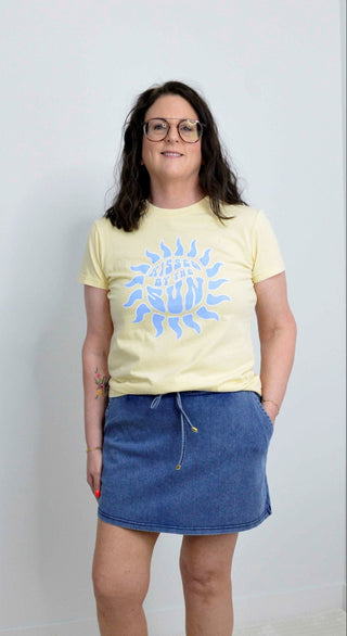 Person wearing a yellow t-shirt with a blue graphic design and a denim skirt on a plain background