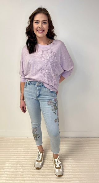 Woman wearing a light lavender blouse and light blue jeans with floral embroidery, standing against a white background.