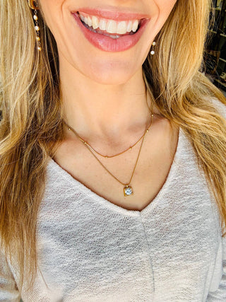 A close-up lifestyle shot of a woman wearing a delicate gold double-layer necklace with a small round clear gemstone pendant, styled with matching gold drop earrings and a light gray knit top.