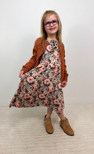 A young girl with glasses is wearing a brown cardigan, a pink and green floral dress, and tan boots, standing against a plain light gray wall and beige carpet.