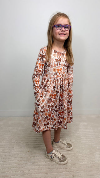 A young girl with long blonde hair and purple-framed glasses stands in a room wearing a long-sleeve midi dress with a brown and orange butterfly pattern on a light pinkish-beige background. She is wearing white sneakers with glittery soles.