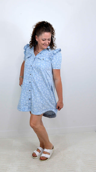 A woman modeling a light blue short-sleeve button-up dress with a white floral pattern, paired with white slide sandals, standing against a plain white wall.