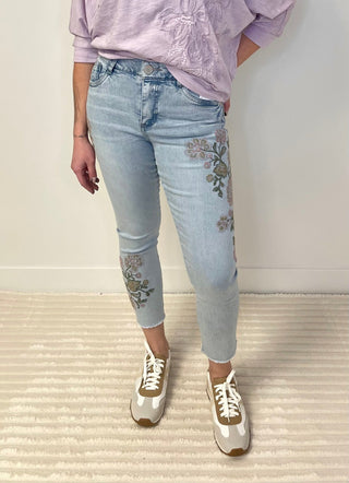 Person wearing light blue jeans with floral embroidery and a lavender top against a plain background