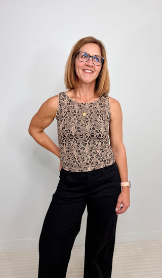 A woman with blonde hair and glasses wearing a sleeveless beige and black patterned top with black pants, standing against a plain light gray wall.