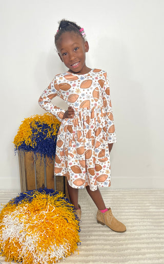 A young girl stands indoors modeling a long-sleeve white dress with an all-over football print. She is posed next to decorative yellow, blue, and white pom-poms against a plain light wall.