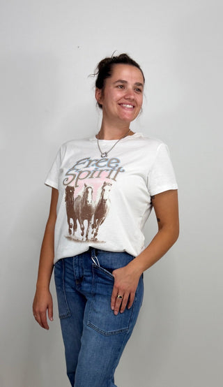 A woman wearing a white graphic t-shirt with a brown llama print and the words 'Free Spirit' in blue jeans, standing against a plain light gray wall.