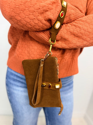 A brown suede crossbody bag with gold-tone hardware and a wide brown strap featuring cream and red oval accents, worn across a person's torso over an orange sweater and blue jeans.