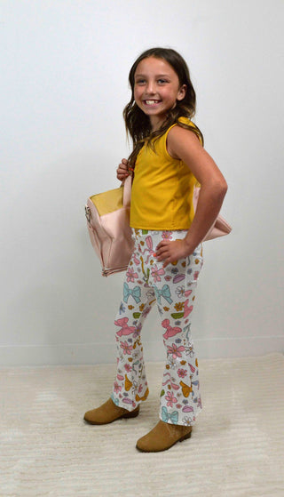 Young girl wearing a yellow top and colorful patterned pants, standing against a plain background.