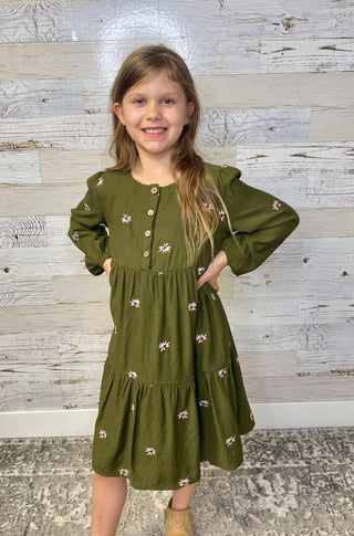 Young girl wearing a green dress with floral patterns against a wooden panel background