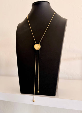 A gold lariat necklace with a round disc pendant and long dangling chains, displayed on a black jewelry bust against a light off-white background.