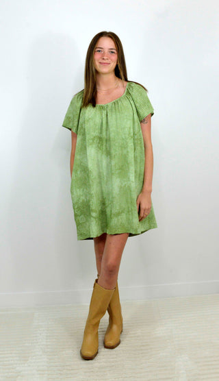 A woman modeling a short-sleeve, knee-length green dress with a textured pattern, paired with tan ankle boots, standing against a plain white wall.