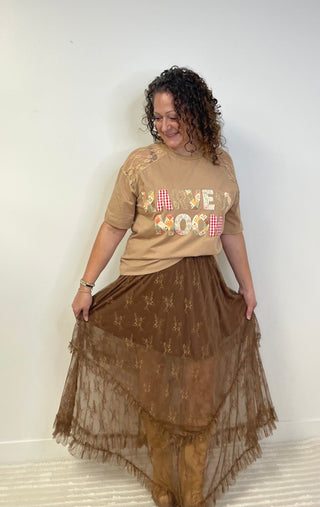 A woman modeling a beige graphic t-shirt with 'Harvest Moon' in colorful letters and a brown sheer embroidered midi skirt, standing against a plain white wall.